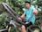 A close-up view of a bicycle chain held by a person in the foreground, with a blurred background of greenery and a bicycle. The chain is metallic and intricate, reflecting the surrounding light.
