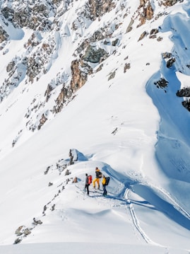 A group of four people wearing colorful winter clothing engage in a mountaineering or skiing adventure on a snow-covered mountain slope. The slope is steep, with visible tracks in the snow indicating their path. Jagged rocks protrude through the snow, adding to the rugged terrain.