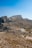 A rugged, rocky landscape stretches across the foreground with a cluster of white, modern buildings situated amid the arid terrain. Sparse vegetation is scattered across the scene, and the land rises to form towering, barren mountains under a clear blue sky.