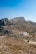 A rugged, rocky landscape stretches across the foreground with a cluster of white, modern buildings situated amid the arid terrain. Sparse vegetation is scattered across the scene, and the land rises to form towering, barren mountains under a clear blue sky.