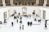 A spacious art gallery with high ceilings and classical architecture is filled with visitors. The gallery features several sculptures on pedestals and paintings hanging on the white walls. People are walking around, some are observing the artworks while others are engaged in conversation. The overall setting is well-lit and sophisticated.