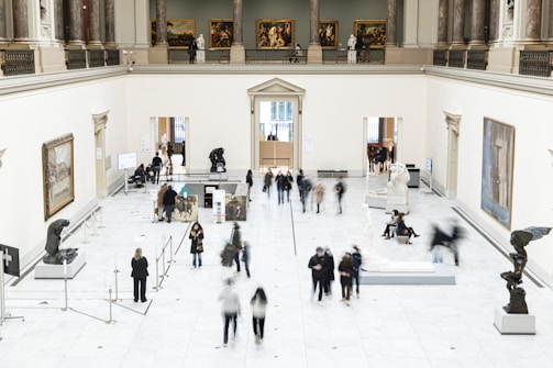 A spacious art gallery with high ceilings and classical architecture is filled with visitors. The gallery features several sculptures on pedestals and paintings hanging on the white walls. People are walking around, some are observing the artworks while others are engaged in conversation. The overall setting is well-lit and sophisticated.