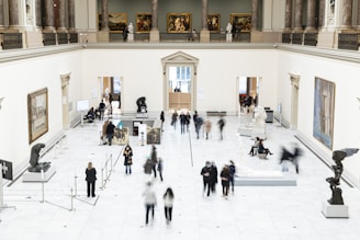 A spacious art gallery with high ceilings and classical architecture is filled with visitors. The gallery features several sculptures on pedestals and paintings hanging on the white walls. People are walking around, some are observing the artworks while others are engaged in conversation. The overall setting is well-lit and sophisticated.