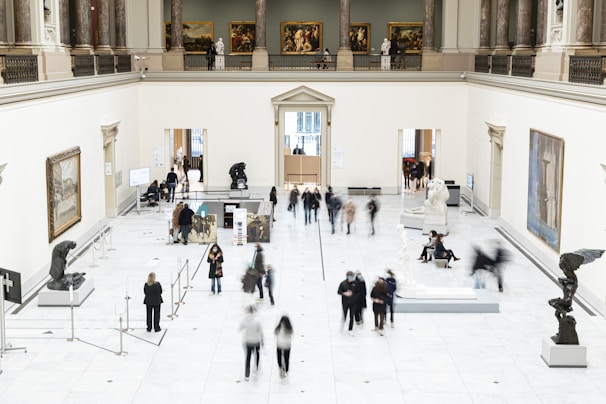 A spacious art gallery with high ceilings and classical architecture is filled with visitors. The gallery features several sculptures on pedestals and paintings hanging on the white walls. People are walking around, some are observing the artworks while others are engaged in conversation. The overall setting is well-lit and sophisticated.
