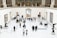 A spacious art gallery with high ceilings and classical architecture is filled with visitors. The gallery features several sculptures on pedestals and paintings hanging on the white walls. People are walking around, some are observing the artworks while others are engaged in conversation. The overall setting is well-lit and sophisticated.