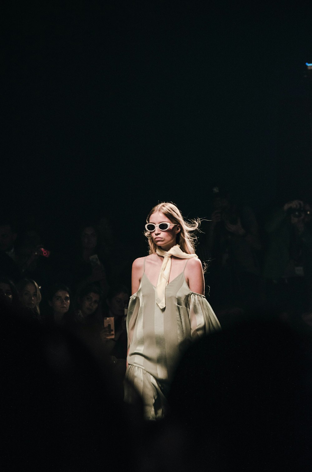 Runway model presenting a pale layered look beneath a single spotlight
