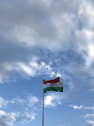 A flagpole with a tricolor flag waving against a backdrop of a partly cloudy sky. The sky features various shades of blue and white, with the flag prominently displaying horizontal stripes of saffron, white, and green.
