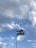 A flagpole with a tricolor flag waving against a backdrop of a partly cloudy sky. The sky features various shades of blue and white, with the flag prominently displaying horizontal stripes of saffron, white, and green.