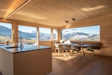 A modern kitchen with wooden walls and ceiling features large windows offering a panoramic view of snow-capped mountains and lush green fields. The kitchen island has a sleek countertop with an integrated sink and faucet. A dining area with minimalist chairs surrounds a wooden dining table. The natural light flooding the room creates an inviting atmosphere.
