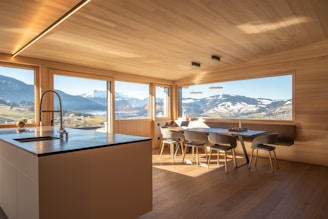 A modern kitchen with wooden walls and ceiling features large windows offering a panoramic view of snow-capped mountains and lush green fields. The kitchen island has a sleek countertop with an integrated sink and faucet. A dining area with minimalist chairs surrounds a wooden dining table. The natural light flooding the room creates an inviting atmosphere.