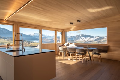 A modern kitchen with wooden walls and ceiling features large windows offering a panoramic view of snow-capped mountains and lush green fields. The kitchen island has a sleek countertop with an integrated sink and faucet. A dining area with minimalist chairs surrounds a wooden dining table. The natural light flooding the room creates an inviting atmosphere.