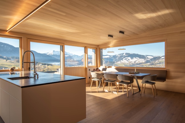 A modern kitchen with wooden walls and ceiling features large windows offering a panoramic view of snow-capped mountains and lush green fields. The kitchen island has a sleek countertop with an integrated sink and faucet. A dining area with minimalist chairs surrounds a wooden dining table. The natural light flooding the room creates an inviting atmosphere.