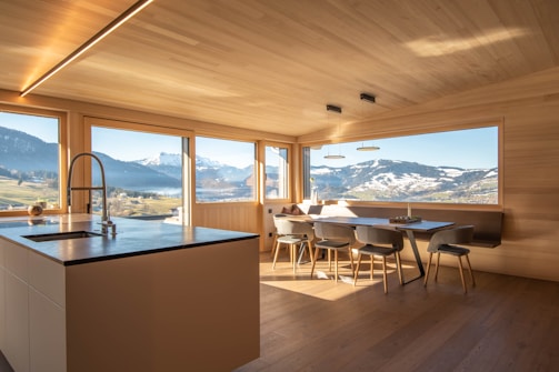 A modern kitchen with wooden walls and ceiling features large windows offering a panoramic view of snow-capped mountains and lush green fields. The kitchen island has a sleek countertop with an integrated sink and faucet. A dining area with minimalist chairs surrounds a wooden dining table. The natural light flooding the room creates an inviting atmosphere.