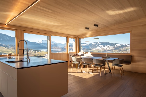 A modern kitchen with wooden walls and ceiling features large windows offering a panoramic view of snow-capped mountains and lush green fields. The kitchen island has a sleek countertop with an integrated sink and faucet. A dining area with minimalist chairs surrounds a wooden dining table. The natural light flooding the room creates an inviting atmosphere.