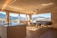 A modern kitchen with wooden walls and ceiling features large windows offering a panoramic view of snow-capped mountains and lush green fields. The kitchen island has a sleek countertop with an integrated sink and faucet. A dining area with minimalist chairs surrounds a wooden dining table. The natural light flooding the room creates an inviting atmosphere.