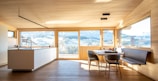 A spacious and modern kitchen interior with large windows providing an expansive view of a scenic landscape. The room features light wooden flooring and ceiling, with a minimalistic countertop and sink. There's a dining area with a table and chairs next to a built-in bench, all bathed in natural light.
