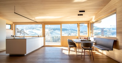 A spacious and modern kitchen interior with large windows providing an expansive view of a scenic landscape. The room features light wooden flooring and ceiling, with a minimalistic countertop and sink. There's a dining area with a table and chairs next to a built-in bench, all bathed in natural light.