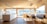 A spacious and modern kitchen interior with large windows providing an expansive view of a scenic landscape. The room features light wooden flooring and ceiling, with a minimalistic countertop and sink. There's a dining area with a table and chairs next to a built-in bench, all bathed in natural light.