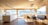 A spacious and modern kitchen interior with large windows providing an expansive view of a scenic landscape. The room features light wooden flooring and ceiling, with a minimalistic countertop and sink. There's a dining area with a table and chairs next to a built-in bench, all bathed in natural light.