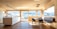 A spacious and modern kitchen interior with large windows providing an expansive view of a scenic landscape. The room features light wooden flooring and ceiling, with a minimalistic countertop and sink. There's a dining area with a table and chairs next to a built-in bench, all bathed in natural light.
