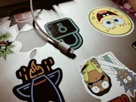 A collection of colorful cartoon character stickers is placed on a silver laptop surface. An assortment of stickers features animated characters and a charging cable draped over the laptop.
