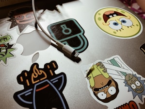 A collection of colorful cartoon character stickers is placed on a silver laptop surface. An assortment of stickers features animated characters and a charging cable draped over the laptop.