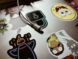 A collection of colorful cartoon character stickers is placed on a silver laptop surface. An assortment of stickers features animated characters and a charging cable draped over the laptop.