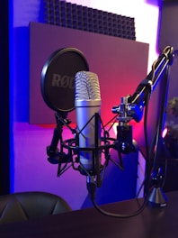 A professional microphone setup is prominently displayed, featuring a silver microphone mounted on a suspension arm with a shock mount. A pop filter is positioned in front of the microphone. The background includes acoustic foam panels on the wall, creating a studio environment. The lighting casts a blend of blue and purple hues across the space.