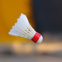 A close-up image of a badminton shuttlecock with white feathery tops and a red base, set against a blurred background that includes shades of yellow and gray.