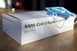 A SARS-CoV-2 rapid antigen test box placed on a wooden surface with a stack of blue surgical masks on top. The lighting casts shadows, indicating a bright environment.