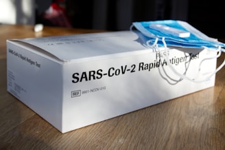 A SARS-CoV-2 rapid antigen test box placed on a wooden surface with a stack of blue surgical masks on top. The lighting casts shadows, indicating a bright environment.