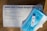 A SARS-CoV-2 rapid antigen test kit is displayed on a wooden surface. The white box includes text detailing the contents and instructions. A blue surgical mask is placed on top of the box alongside a white test device.