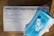 A SARS-CoV-2 rapid antigen test kit is displayed on a wooden surface. The white box includes text detailing the contents and instructions. A blue surgical mask is placed on top of the box alongside a white test device.