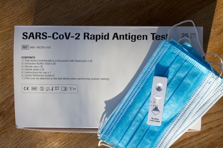 A SARS-CoV-2 rapid antigen test kit is displayed on a wooden surface. The white box includes text detailing the contents and instructions. A blue surgical mask is placed on top of the box alongside a white test device.