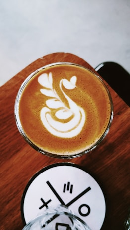 A cup of coffee with intricate latte art consisting of a white pattern resembling leaves and hearts on a brown surface. The drink is placed on a wooden table, accompanied by a white coaster with a modern black design. The background features a soft gray tone.