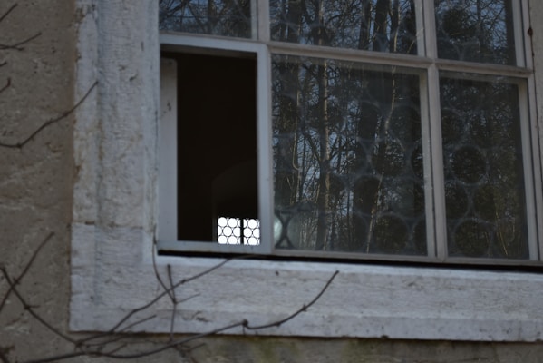 A section of an old window with multiple panes features glass with circular patterns. The window is set in a weathered, beige wall. Outside, the glass reflects a view of trees, while one pane is missing, creating a small opening.