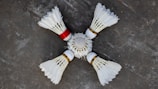 Four white shuttlecocks with feathers arranged in a circular pattern on a grey surface. One shuttlecock has a red band, contrasting with the white feathers and cork tips.