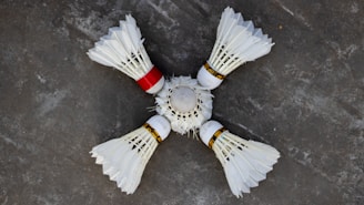 Four white shuttlecocks with feathers arranged in a circular pattern on a grey surface. One shuttlecock has a red band, contrasting with the white feathers and cork tips.