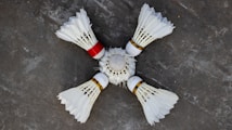 Four white shuttlecocks with feathers arranged in a circular pattern on a grey surface. One shuttlecock has a red band, contrasting with the white feathers and cork tips.