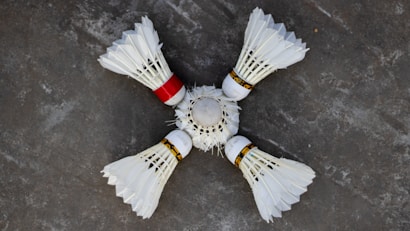 Four white shuttlecocks with feathers arranged in a circular pattern on a grey surface. One shuttlecock has a red band, contrasting with the white feathers and cork tips.