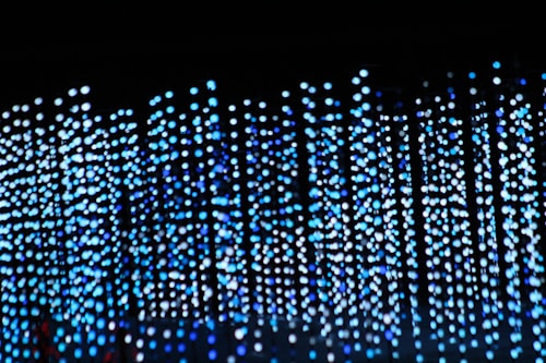 Hundreds of small, blue and white LED lights are suspended in rows against a dark background, creating a cascading waterfall effect. The lights appear blurred, contributing to an abstract and ethereal appearance.