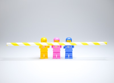 Three colorful toy figures stand in a line, each holding a yellow and white striped barrier. The figures are dressed in space-themed outfits with a simplistic, block-like appearance. The background is a plain light gray, emphasizing the vibrant colors of the toys.