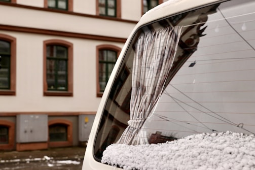 A close-up view of the back window of a white vehicle, partially covered with snow. The window has a curtain, and the reflection of a building with arched windows is visible on the glass. In the background, a multi-story building with brown and white detailing is seen.