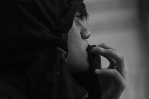 A person is viewed in profile, wearing a dark hooded jacket, seemingly lost in thought. The hand is raised, partially covering the mouth, adding a contemplative element to the scene. The image is captured in black and white, enhancing the focus on the person's expression.