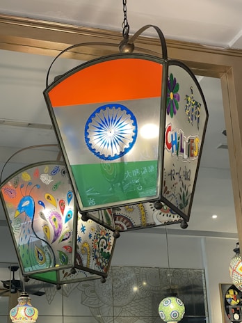 A colorful lantern with intricate designs, featuring an Indian flag motif on one side and vibrant peacock illustrations on another. The lantern is suspended from the ceiling and casts a warm, inviting light. Surrounding the lantern are additional decorative elements with vivid patterns and a mirrored surface reflecting parts of the room.