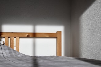 A wooden bed frame catches sunlight, casting shadows onto a plain white wall. The bed has a checkered grey duvet. The corner of the room is visible with stark contrasts between light and shade.