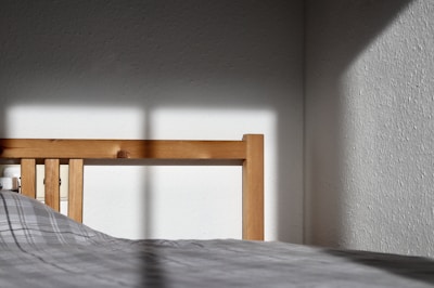 A wooden bed frame catches sunlight, casting shadows onto a plain white wall. The bed has a checkered grey duvet. The corner of the room is visible with stark contrasts between light and shade.