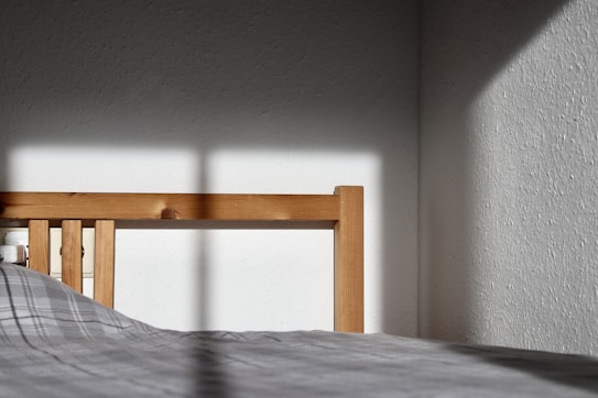 A wooden bed frame catches sunlight, casting shadows onto a plain white wall. The bed has a checkered grey duvet. The corner of the room is visible with stark contrasts between light and shade.