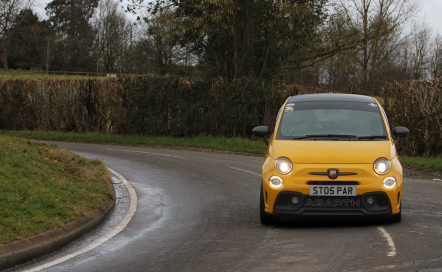 A bright yellow car is driving along a curved, wet road with dense trees and hedges lining the sides. The car is small and compact, with distinctive round headlights and a black roof. Overcast skies suggest it's either early morning or late afternoon.
