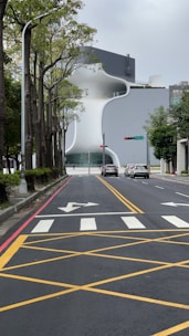 A modern architectural building with a unique curved design stands at the end of a straight road. The road is marked with yellow lines and a pedestrian crosswalk. Trees line the left side of the road, and a few cars are visible in front of the building. Traffic lights and street signs are present, contributing to an urban setting.